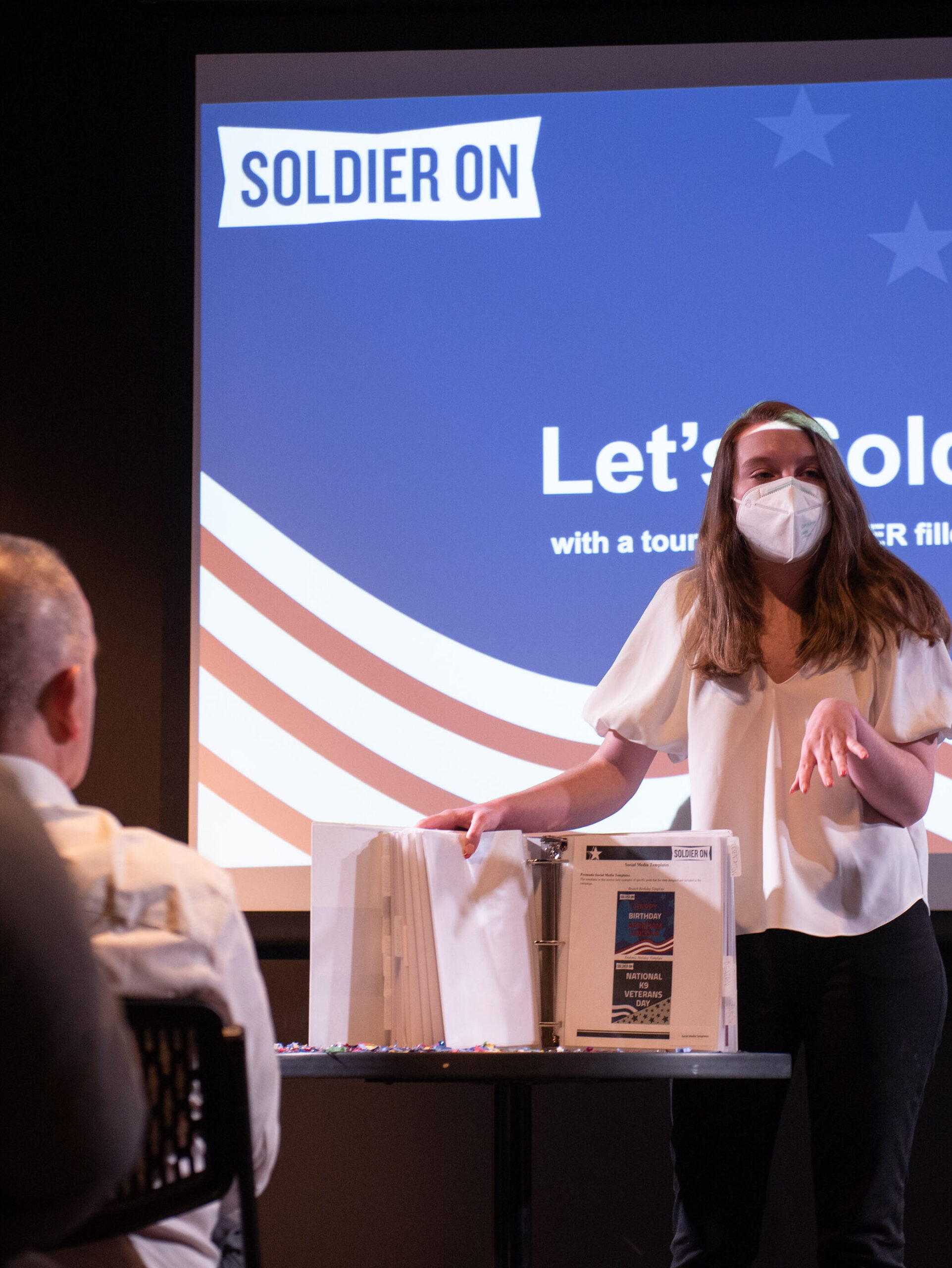 Young woman in face mask shows portfolio book in front of screen with Soldier On logo