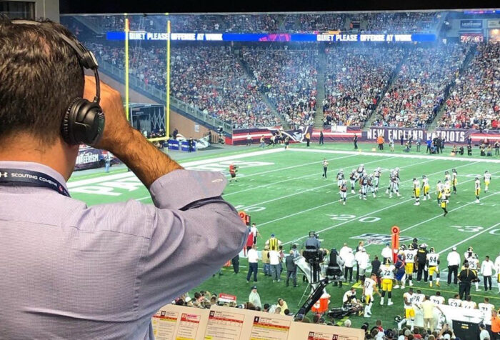 man wearing headphones looks down at game happening on football field from behind glass