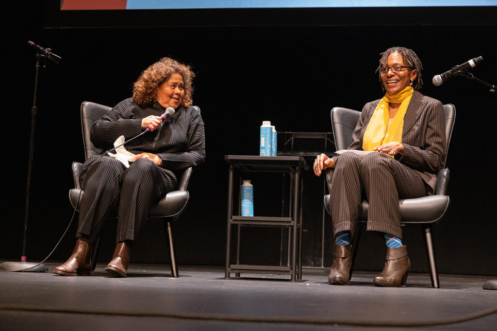 two Black women sit in chairs on stage, speaking into mics, a table with boxed water between them