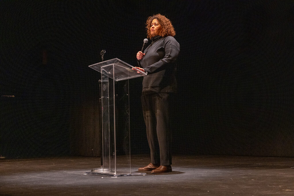 Black woman dressed in Black suit stands on stage behind a clear podium
