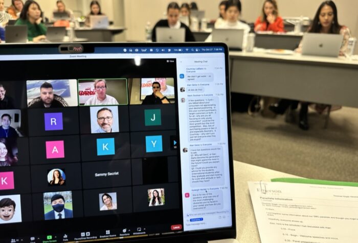 shot of a zoom meeting on a computer screen with students in desks in the background