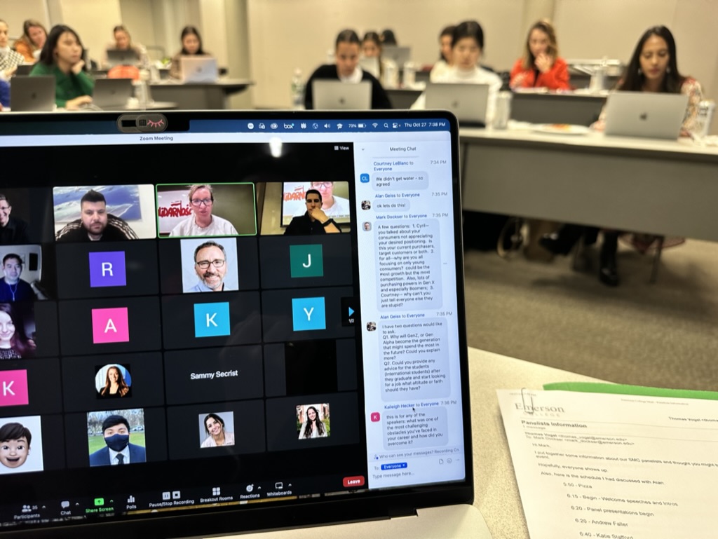 A laptop screen displays a grid view of a the Zoom video call, featuring on-screen SMC alumni and also muted-screen of current students listening to alumni over Zoom inside the Marketing Communication graduate classroom. 