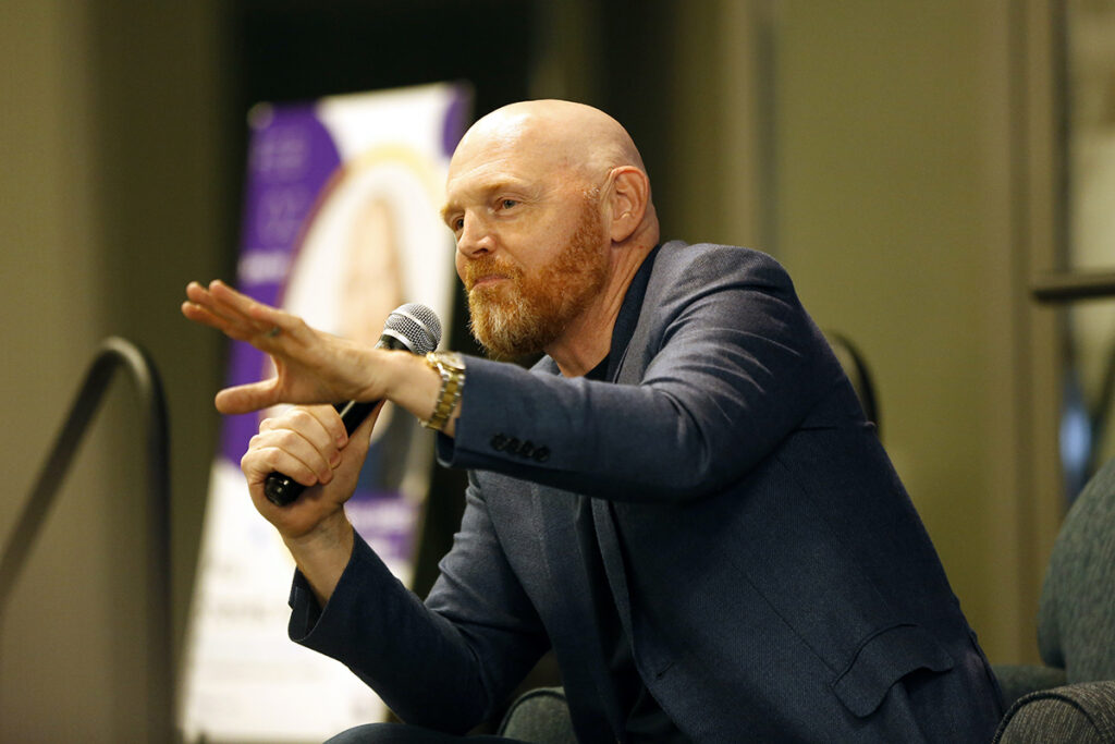 Bill Burr in blue jacket leans forward in chair holding microphone, gesturing with left hand