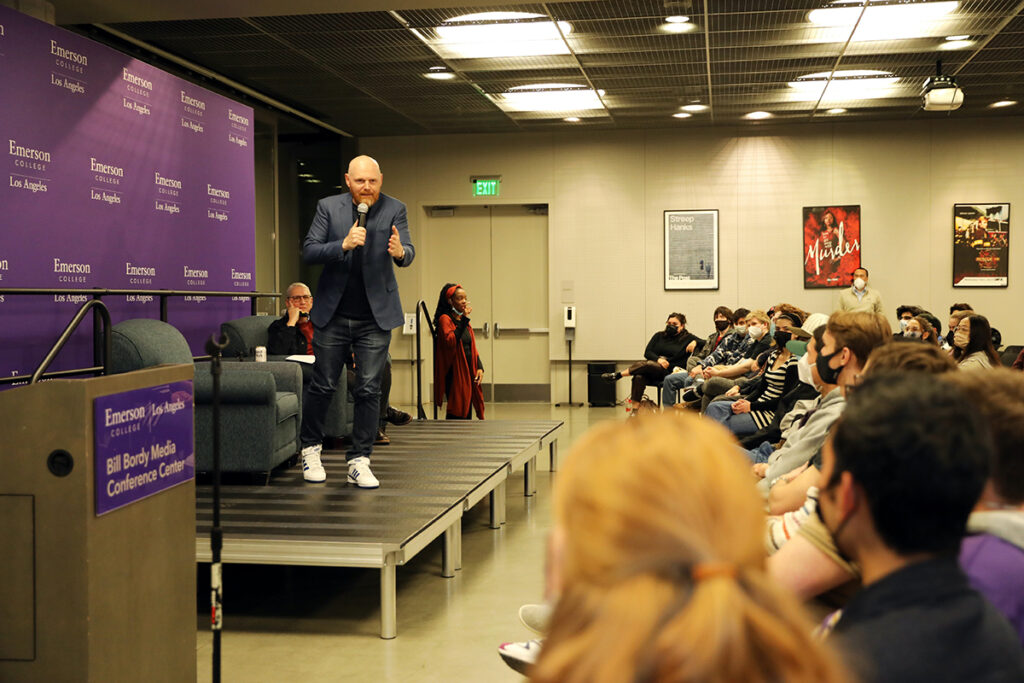 Bill Burr stands on low stage holding mic and gesturing to students in audience. Purple Emerson branded display backs stage, Doug Herzog looks on