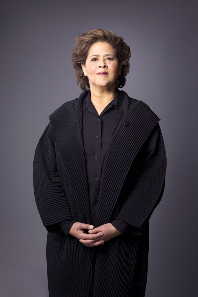 Anna Deavere Smith stands with hands clasped, wearing all black against grey background