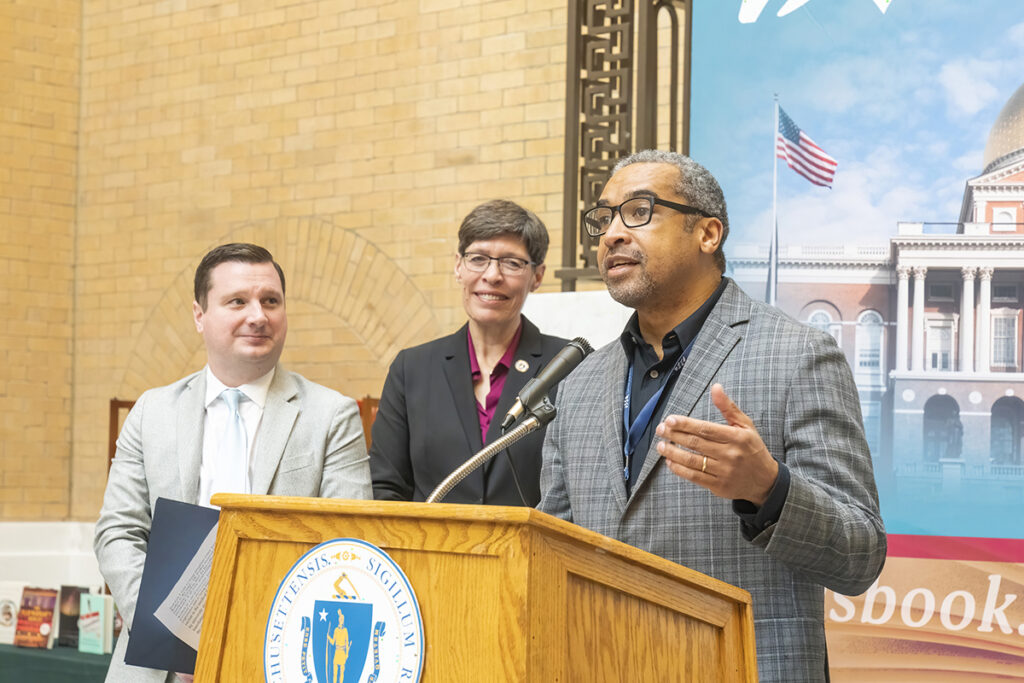 Jerald Walker in grey suit jacket speaks behind podium with Massachusetts state seal, painting of state house behind him. Man and woman look on