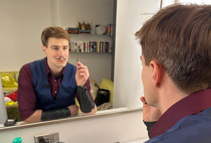 man in red shirt and blue vest looks in mirror as he draws on a fake mustache