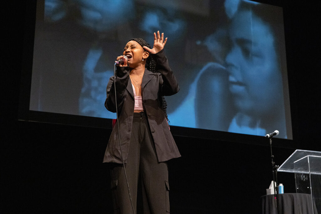 Young Black woman in brown suite sings into mic as images of Civil Rights movement projected onto screen in background