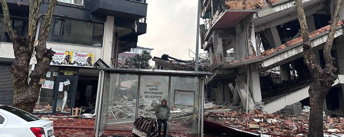 man stands at bus stop framed by rubble an partially collapsed buildings