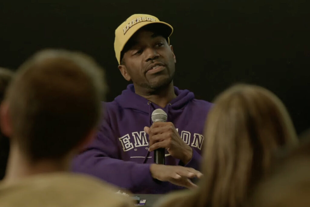 Black man in purple Emerson sweatshirt, yellow Emerson baseball hat speaks into mic, heads of audience members visible in front