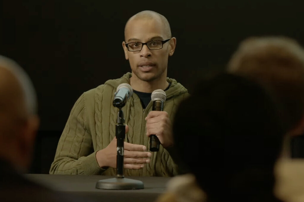 Black man with shaved head in glasses, olive green sweater, speaks into mic while gesturing, heads of audience members visible in front
