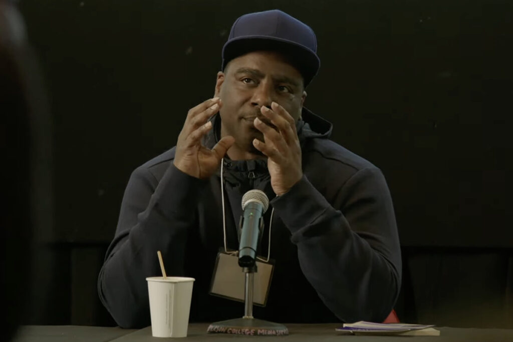 Black man wearing dark sweatshirt, blue baseball hat, gestures behind mic with paper cup in front of him