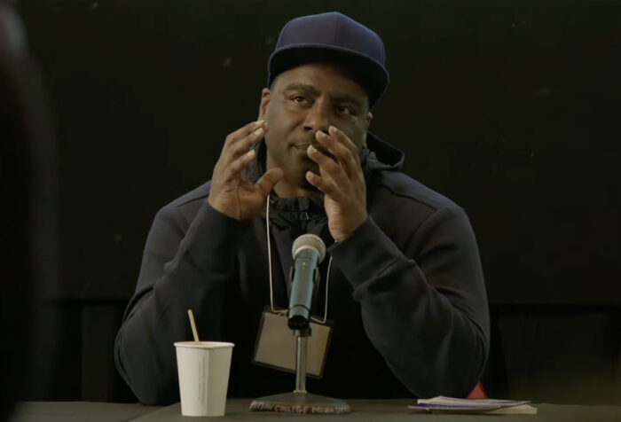 man in black sweatshirt, blue baseball hat gestures and speaks into mic