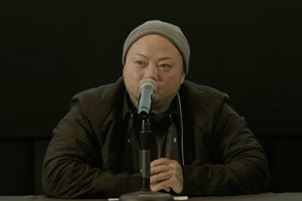 Asian man wearing knit cap, dark jacket, speaks into mic