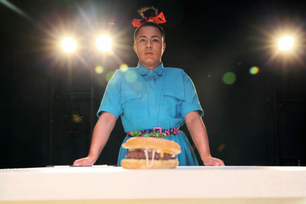 Trans actor in blue dress, hair in topknot, stands behind table on which rests a hamburger