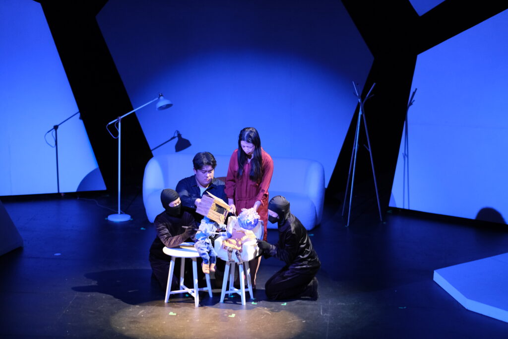 On stage, Yuning Su sits while Wisteria Deng stands, and two puppeteers work two puppets