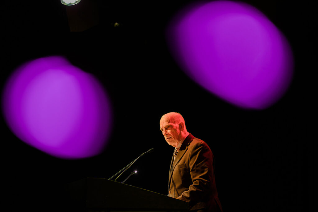 Bill Gilligan speaks at podium on darkened stage with purple lights behind him