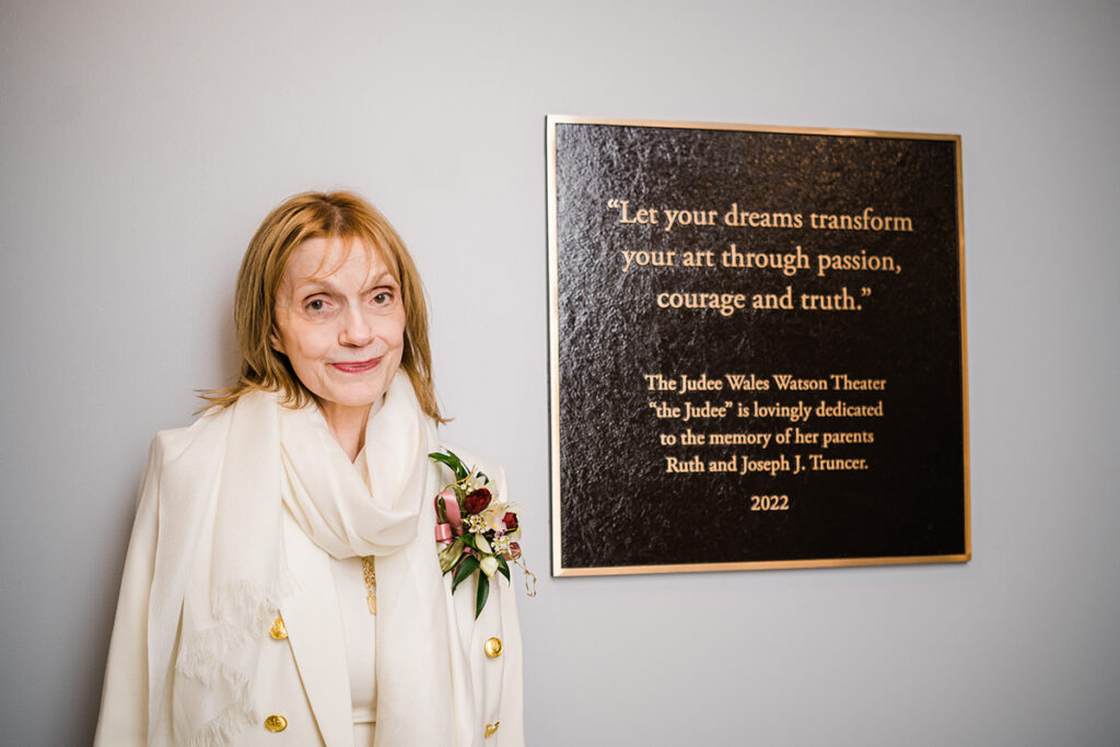 Judee Wales Watson, in cream colored suite and corsage, stands next to plaque that reads, "Let your dreams transform your art through passion courage and truth. The Judee Wales Watson Theater 'the Judee' is lovingly dedicated to the memory of her parents Ruth and Joseph J. Truncer. 2022"