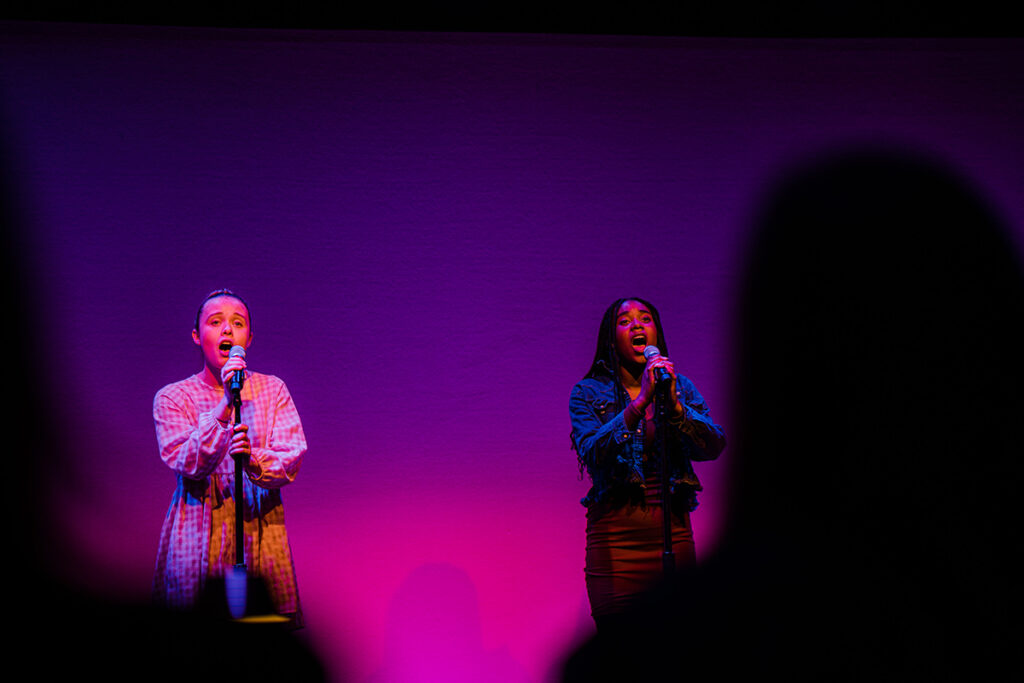 Two young women sing into mics on darkened stage, purple spotlight behind them