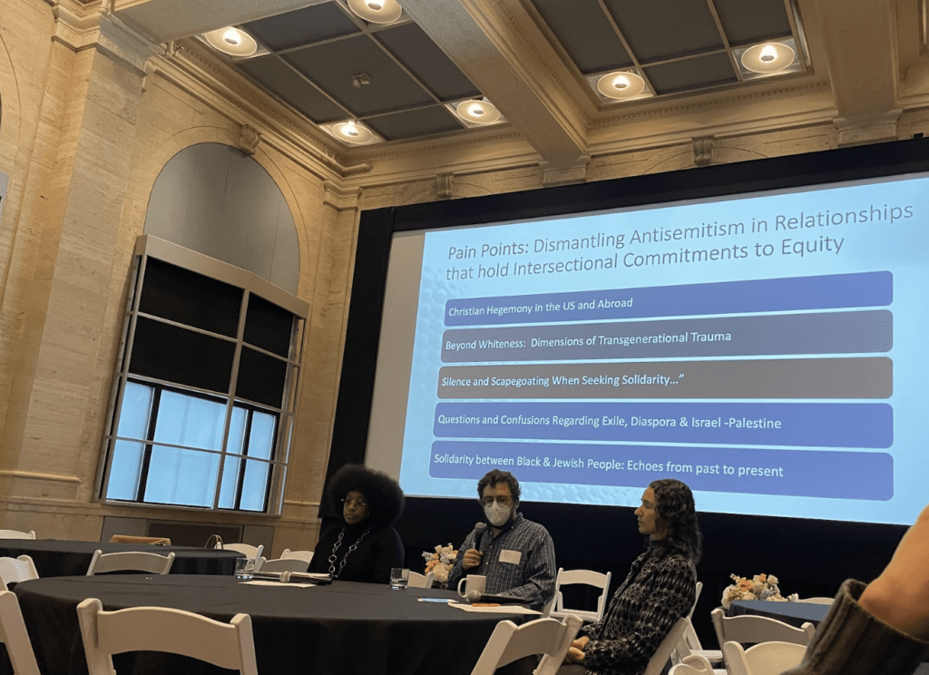 Three people sit at a table with a large projector screen behind them