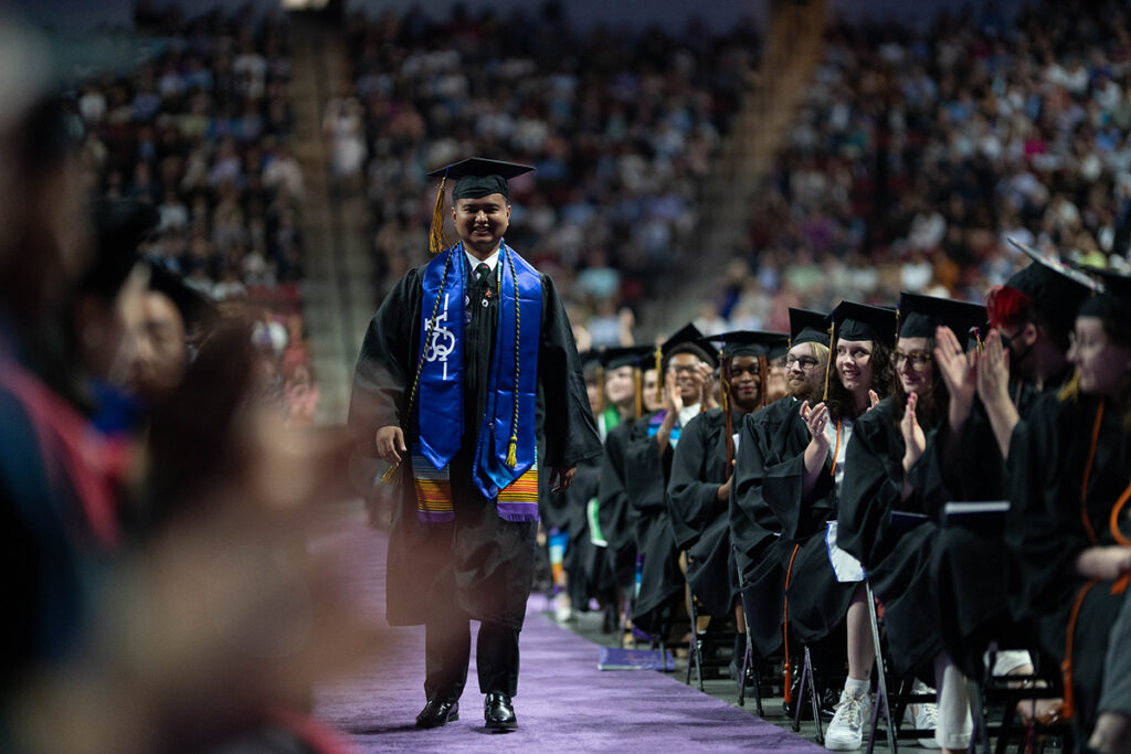 graduate wearing multiple stoles walks down aisle as classmates clap
