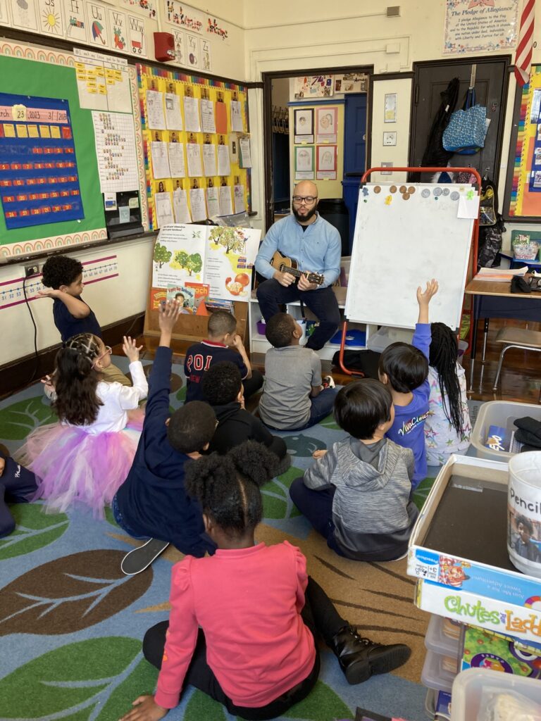 A Boston Music Project professional sits in a chair with a small string instrument with students sitting on the clasroom rug 