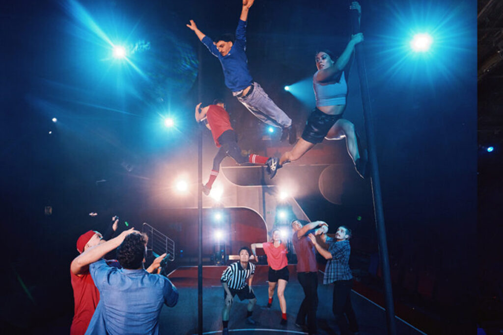 Two acrobats swing from two upright poles as a third dives over their feet, six acrobats on ground look up