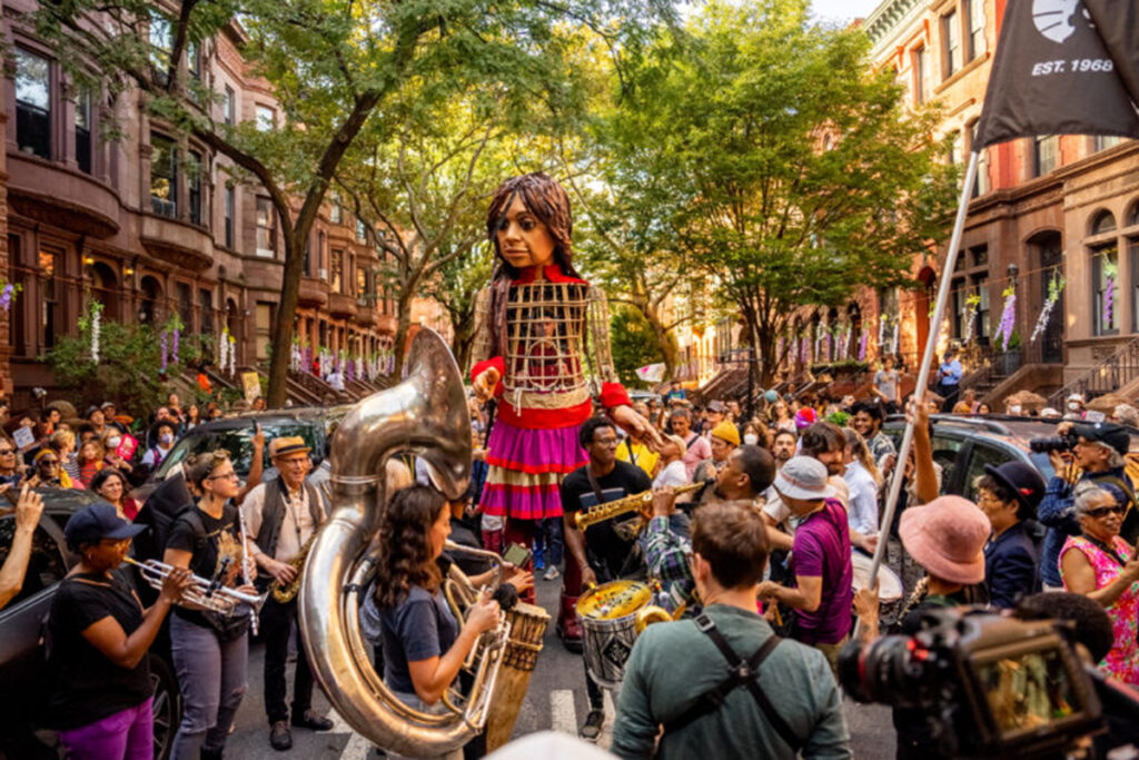 12-foot puppet of Syrian girl in pink and purple dress walks down a Harlem street surrounded by a brass band, spectators