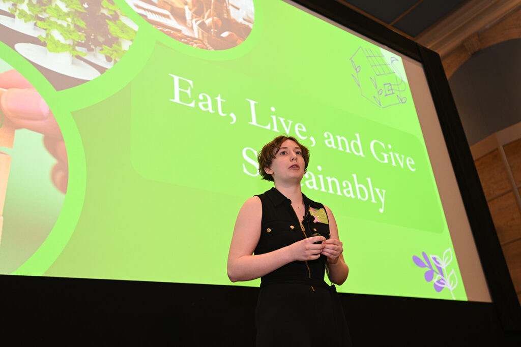 Indigo stands in front of screen with green graphic reading "eat, live and give sustainably"