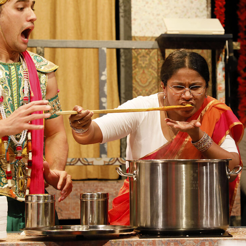 Middle-aged woman in sari tastes something from a pot on the stove as man in traditional Indian dress looks at her, mouth agape