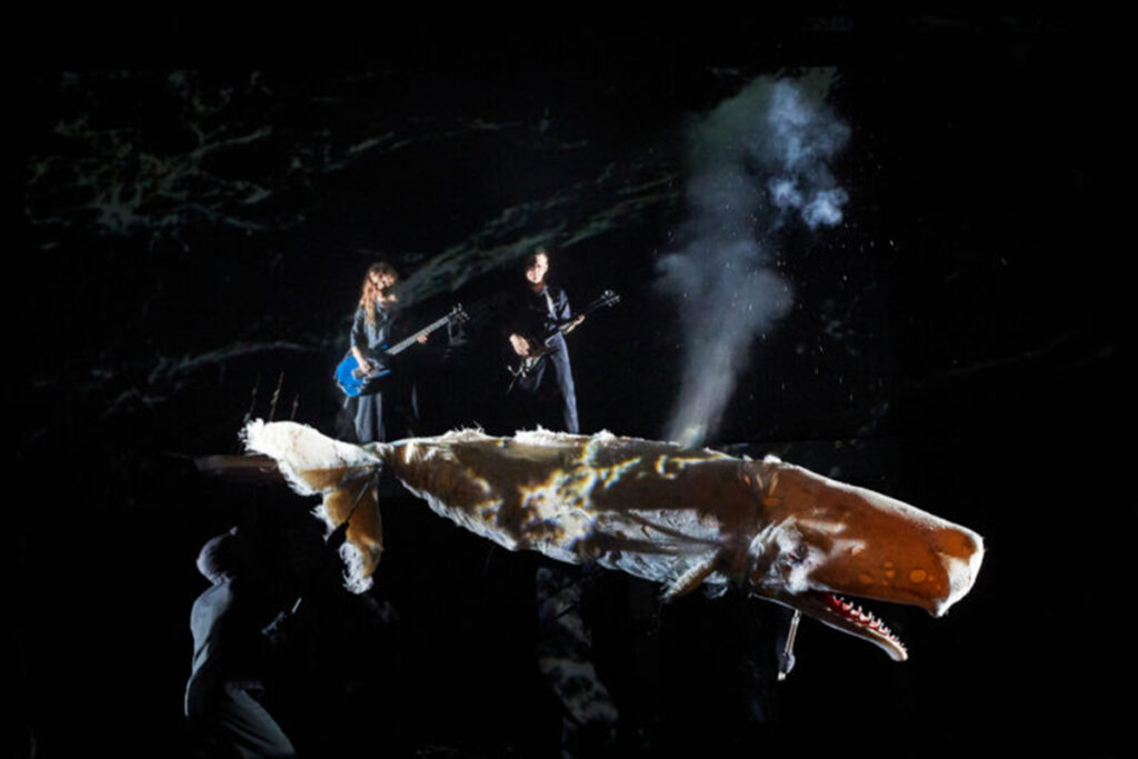 woman and man playing electric guitars appear to stand on top of puppet of sperm whale, vapor coming from blowhole