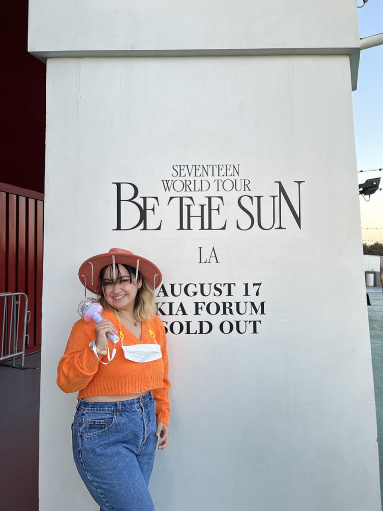 Monica in wide brimmed hat, holding fake plastic microphone in front of sign that reads "Seventeen World Tour Be the Sun LA"