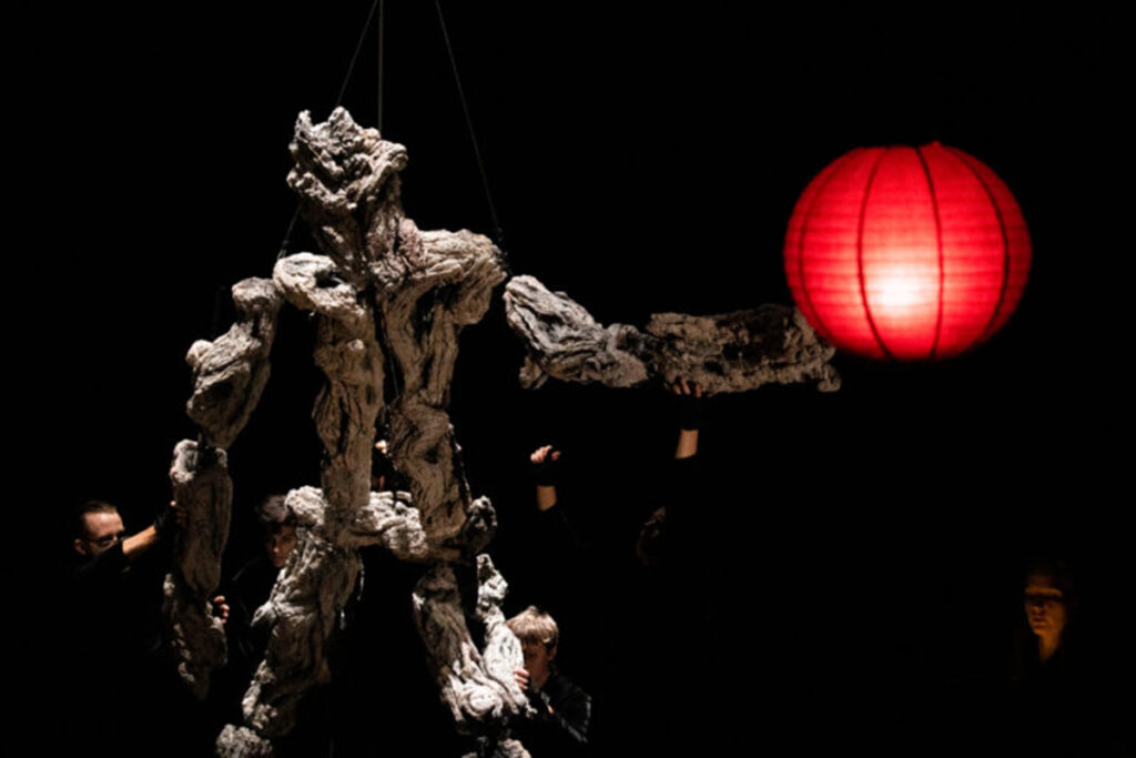 large puppet of a creature appearing made of driftwood stands before a red Chinese lantern