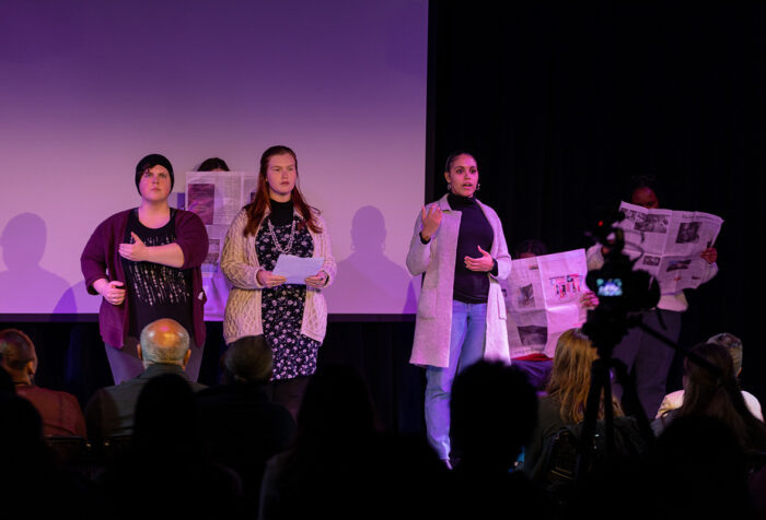 Three people stand on stage with one person in the background with a newspaper in front of their face