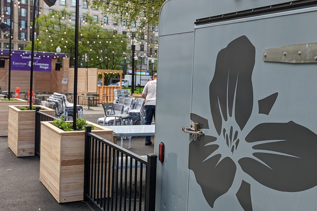 light grey beer truck with dark grey trillium flower painted on side, wooden planters and outdoor stage in background.