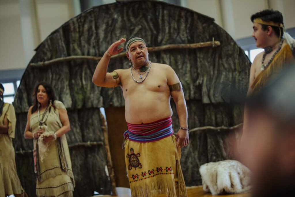 man in traditional Wampanoag dress stands in front of facade of wigwam, other people in traditional dress