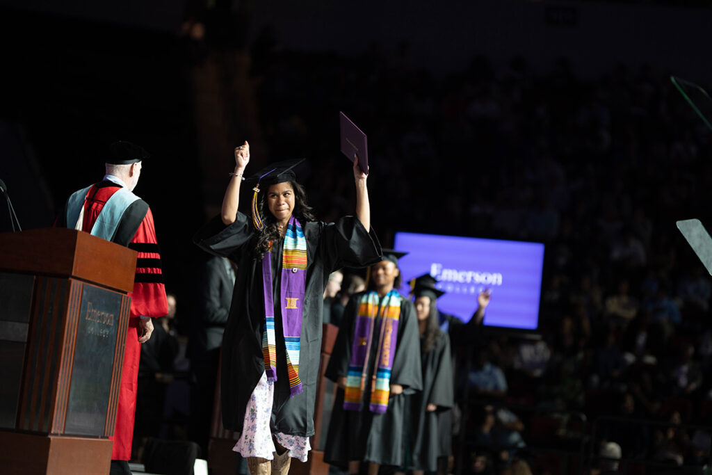 Woman in multicultural stole raises hands, clasping degree cover