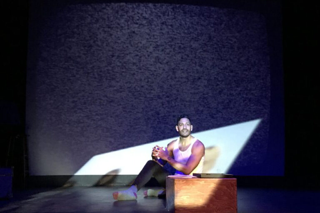 man in white tank top sits on stage floor next to wooden box in a parabola of light