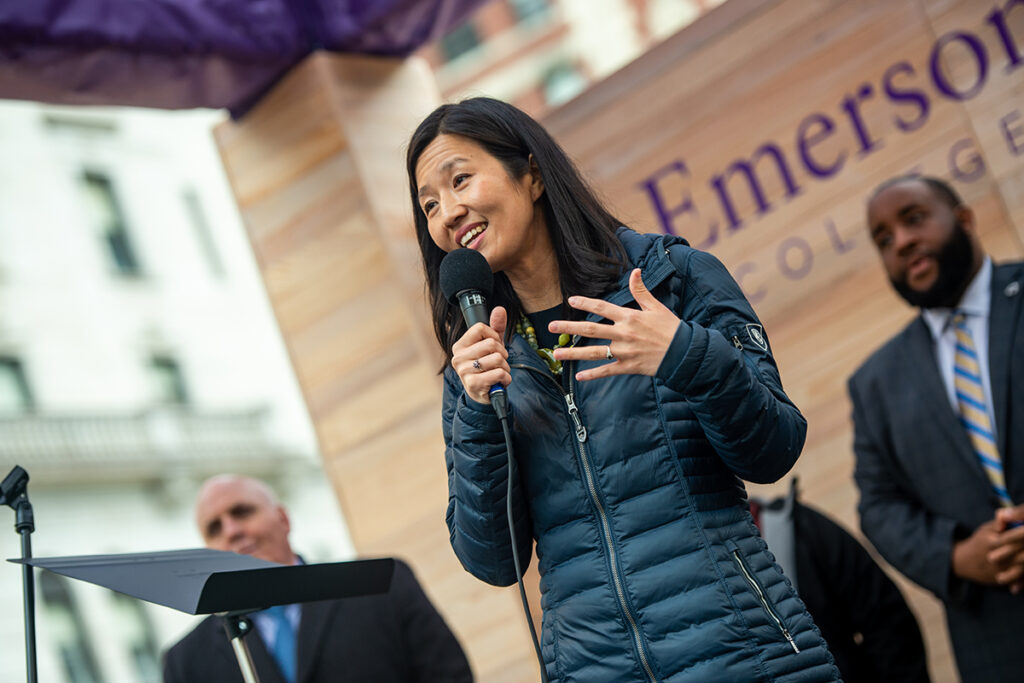 michelle wu, in parka, speaks into microphone, men in suits and Emerson logo visible on wood paneling behind her