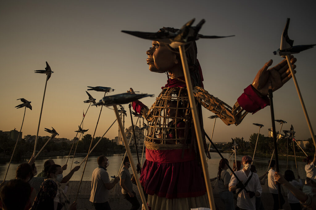 Giant puppet of little girl walks among people holding bird puppets on sticks at dusk