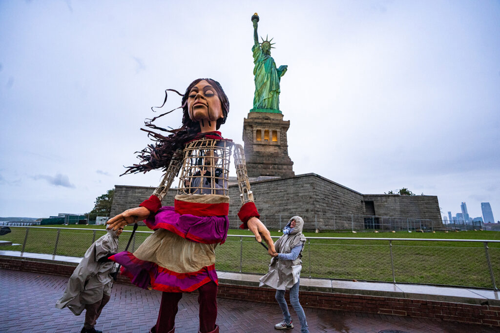 Giant puppet of little girl is maneuvered by two puppeteers in front of the Statue of Liberty