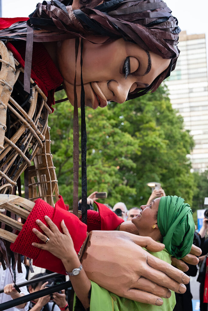 Giant puppet of little girl bends down to rest hands on should of woman in green headscarf.
