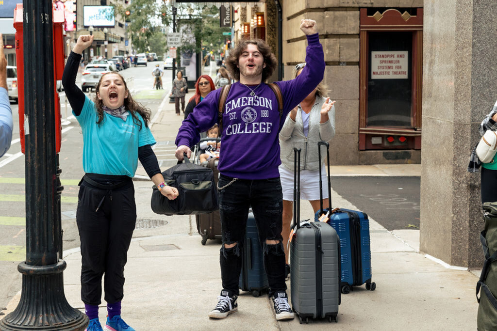 student in blue t shirt and new student in purple Emerson sweatshirt, luggage beside him, raise fists in air
