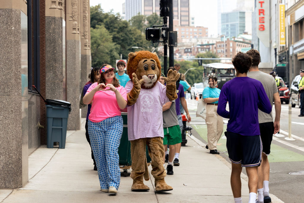 Griff the lion walks down Tremont Street with student in pink t shirt making heart sign with hands