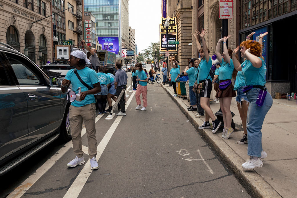 students in blue t-shirts line Tremont Street and cheer for line of cars carrying new students and families