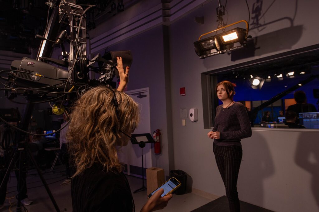 A person raises their hand off camera while another person waits to report in front of the camera