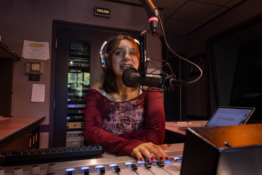 A woman talks in front of a radio microphone with sound board in front of her
