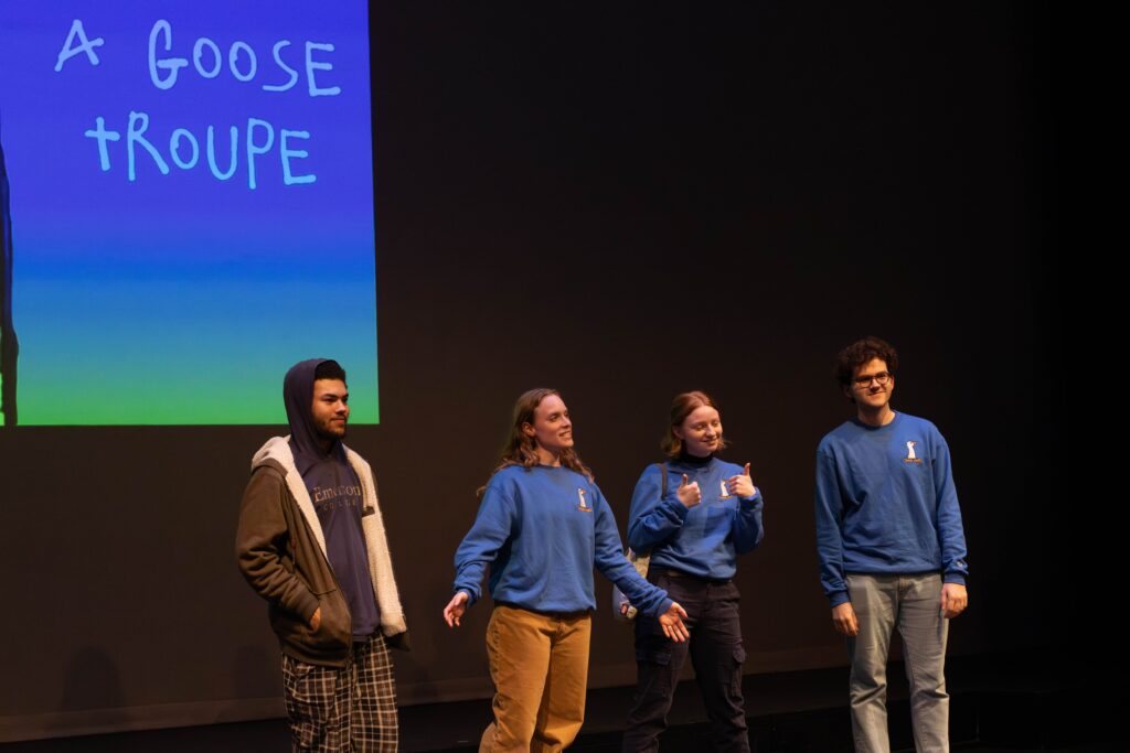 Four people on stage with a projection of the group's name A Goose Troupe