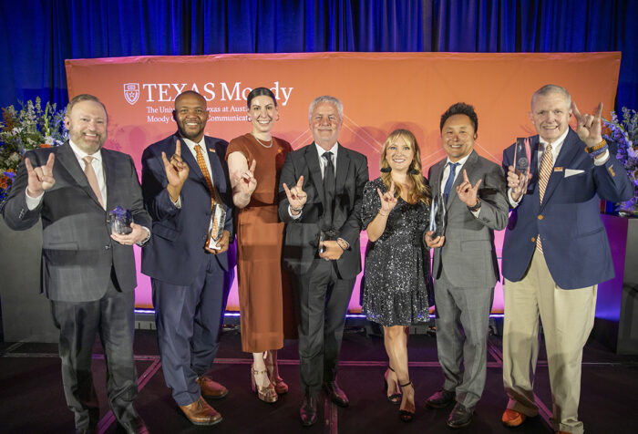 Seven people stand in front of step and repeat, making UT hand sign.