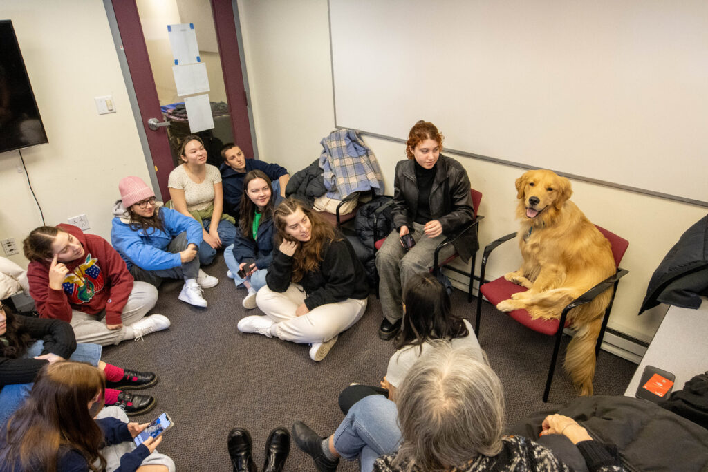 Students sit on the floor and in chairs while Journey the dog sits on a chair
